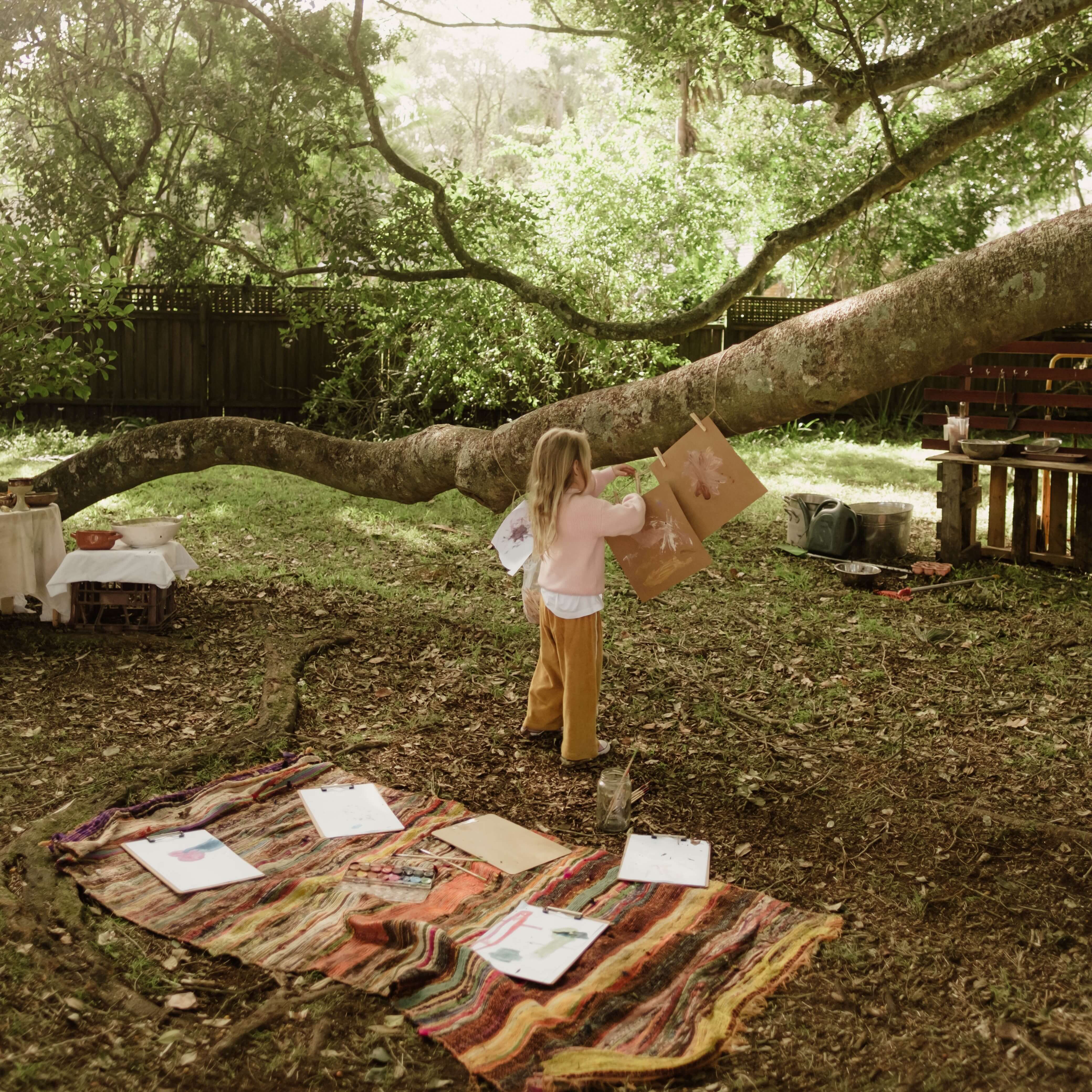A young girl in a backyard, hanging a piece of parchment paper from a clothes line that is attached to a low hanging tree. On the ground, there's a striped colourful blanket with various artworks, paintbrushes and paint.